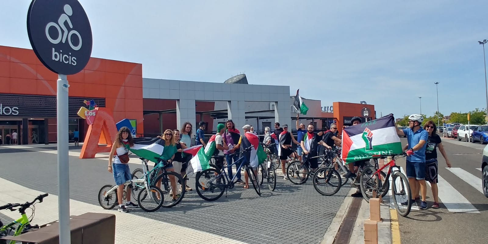 Fotografía del final de la bicicletada frente al centro comercial Alfares en donde se encuentra el Carrefour de Talavera de la Reina con muches de les corredores mostrando banderas palestinas.